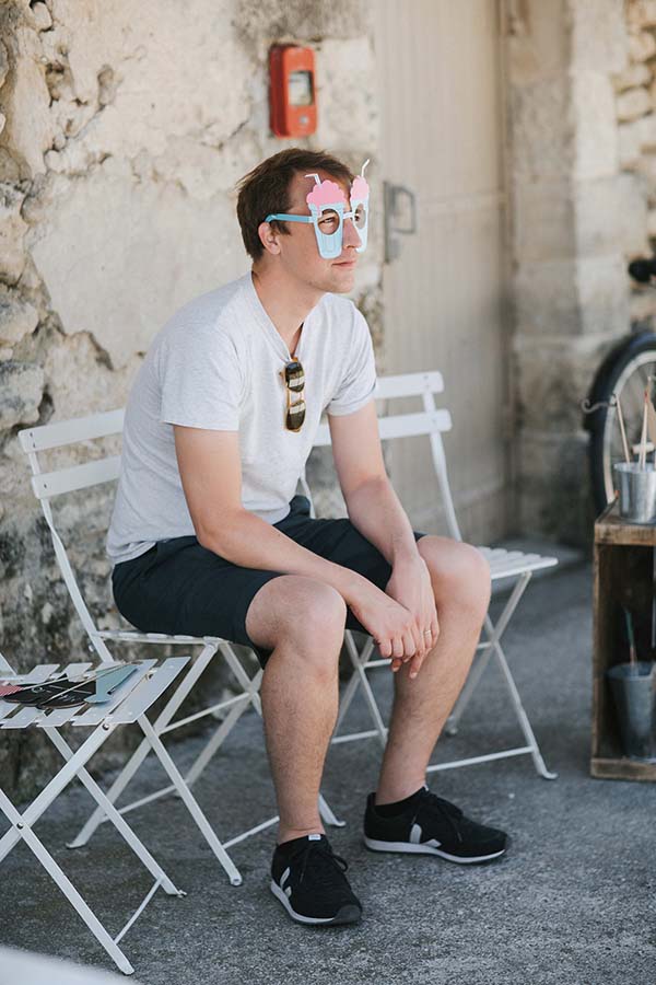 Un homme en t-shirt blanc et short noir portant un masque fantaisie, assis sur une chaise blanche en extérieur.