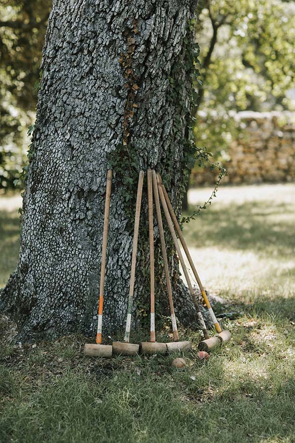Plusieurs crosses de croquet en bois appuyées contre le tronc d'un grand arbre dans un jardin ensoleillé.