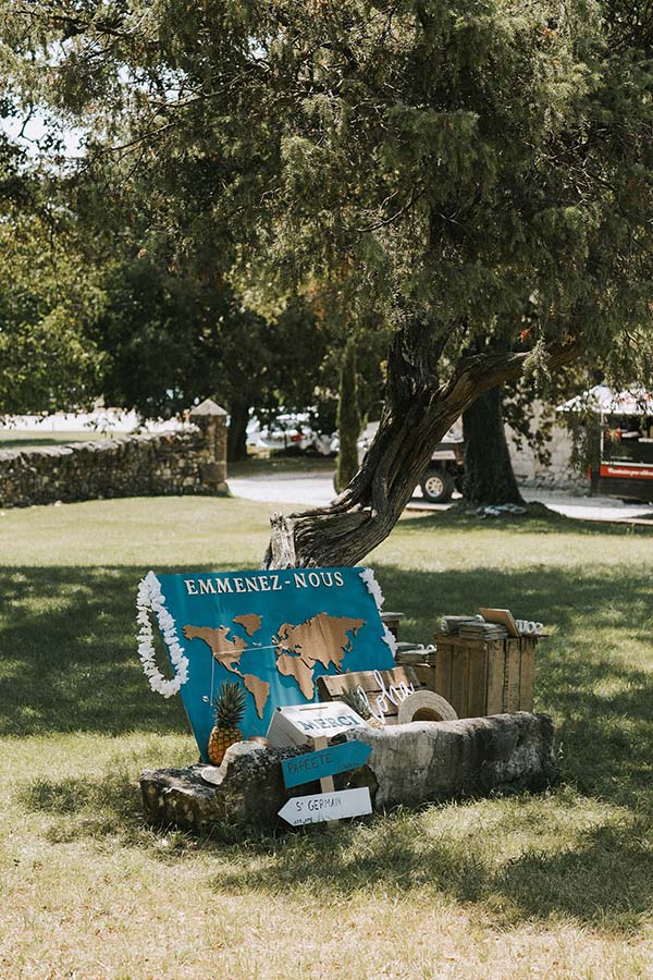 Décoration de mariage originale avec une carte du monde bleue et des panneaux indicateurs sous un grand arbre.