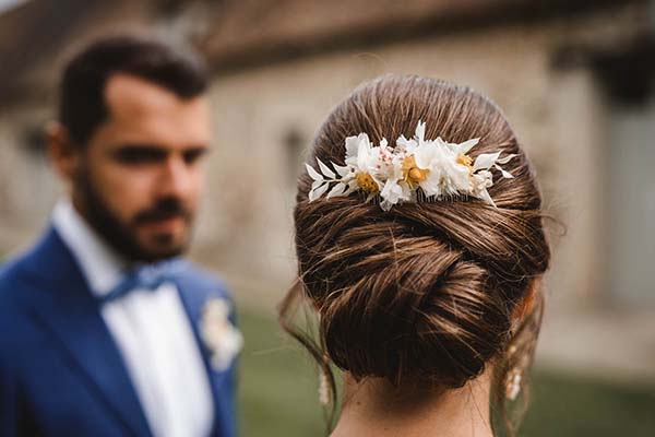 Vue de dos d'une mariée au chignon orné de fleurs blanches, face à son futur époux en costume bleu, ambiance champêtre.