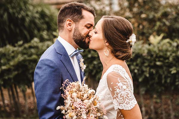 Jeunes mariés s'embrassant tendrement en extérieur, avec un bouquet champêtre et un costume bleu élégant.