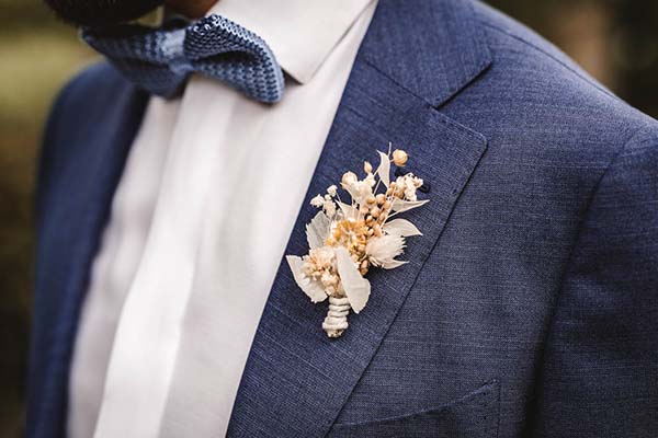 Gros plan d'un marié en costume bleu avec nœud papillon et boutonnière de fleurs séchées pour un style champêtre.