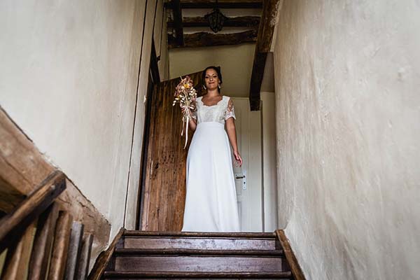 Une mariée en robe blanche tenant un bouquet champêtre, debout en haut d'un escalier en bois dans un intérieur rustique.