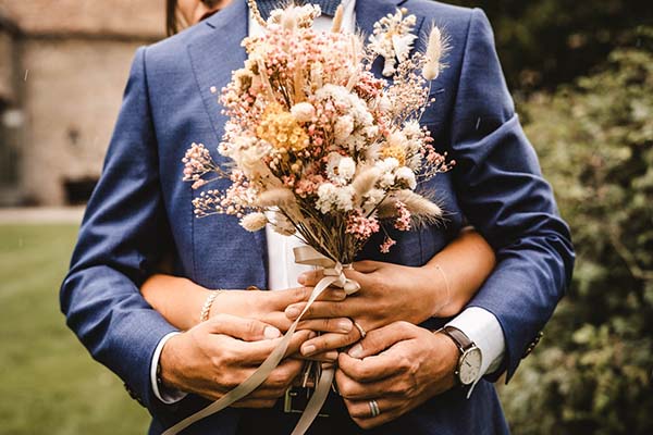 Couple s'enlaçant et tenant un bouquet de fleurs séchées champêtres, le marié portant un costume bleu élégant.