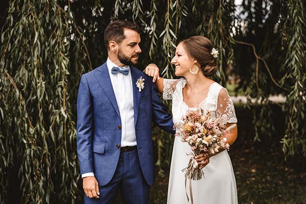 Jeunes mariés complices en tenue élégante posant devant un saule pleureur pour un mariage champêtre et authentique.