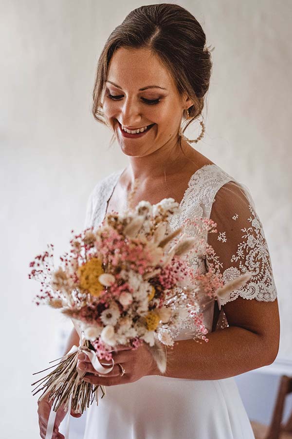 Une mariée souriante en robe blanche tenant un bouquet de fleurs séchées aux tons pastel et naturels.