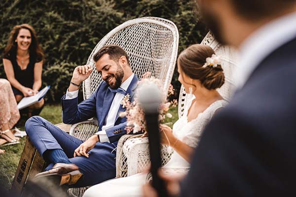 Jeunes mariés souriants installés dans des fauteuils en osier blanc lors d'une cérémonie champêtre en plein air.