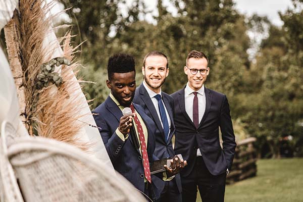Trois hommes en costumes élégants sourient lors d'un mariage champêtre entourés de verdure et de décorations bohèmes.