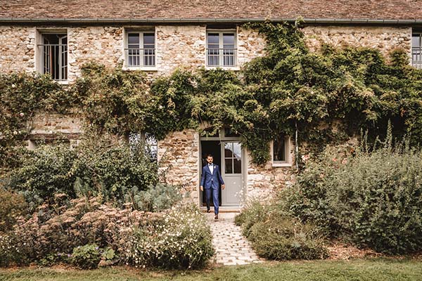 Un marié en costume bleu sort d'une bâtisse en pierre ancienne entourée de végétation luxuriante et champêtre.