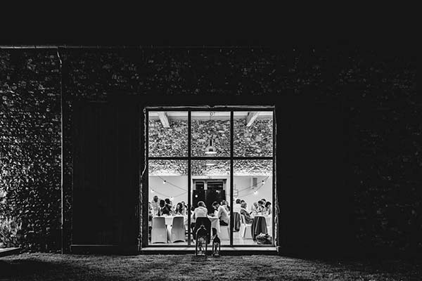 Vue nocturne en noir et blanc d'un dîner de mariage chaleureux à travers une grande baie vitrée entourée de végétation.