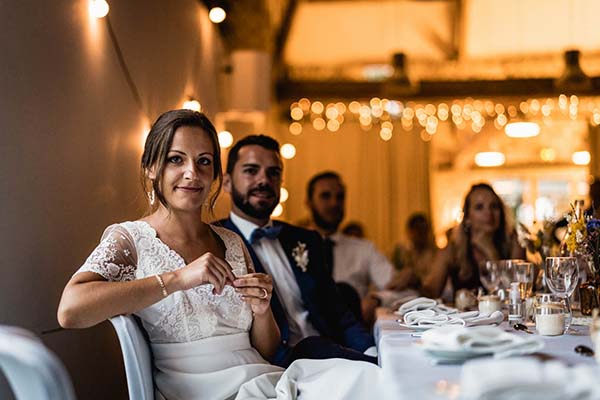 Jeunes mariés souriants à leur table de réception, ambiance chaleureuse et lumineuse avec guirlandes scintillantes.