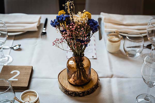 Centre de table champêtre avec fleurs séchées colorées dans un vase ambré sur une table de mariage blanche.