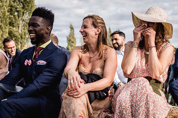Trois invités rient avec complicité lors d'un mariage champêtre, vêtus de tenues élégantes et estivales.