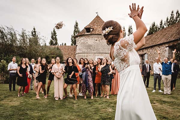 Une mariée en robe blanche lance son bouquet à ses invitées devant un château rustique lors d'un mariage champêtre.