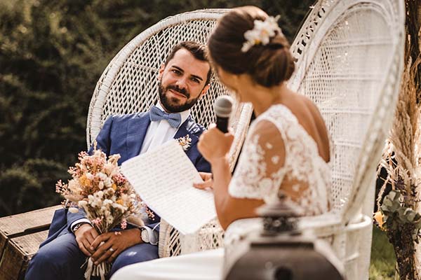 Une mariée lit ses vœux au micro à son futur époux assis dans un fauteuil en osier lors d'un mariage champêtre.