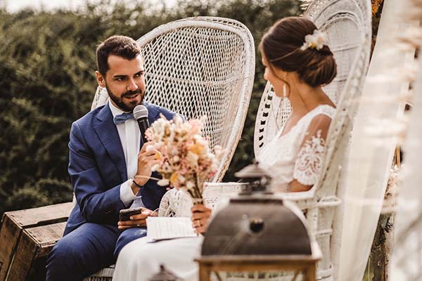 Jeunes mariés en costume bleu et robe blanche discutant dans des fauteuils en osier blanc lors d'un mariage champêtre.