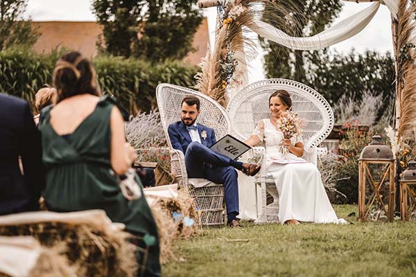 Jeunes mariés assis dans des fauteuils en osier blanc lors d'une cérémonie champêtre et naturelle en plein air.