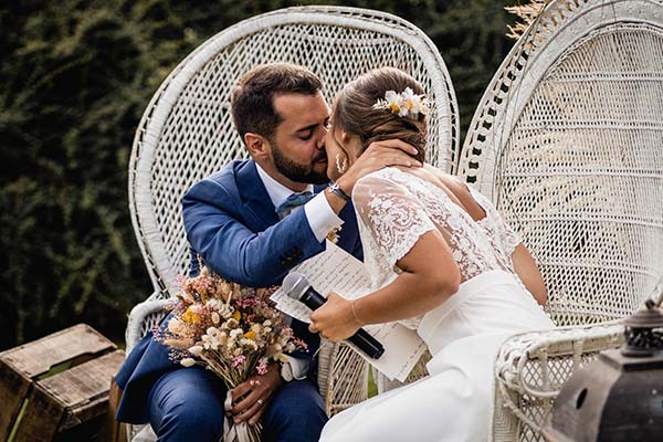 Jeunes mariés s'embrassant tendrement dans des fauteuils en osier blanc lors d'un mariage champêtre et lumineux.