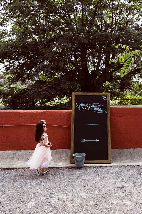 Une petite fille en robe rose poudrée devant un panneau de pétanque, dans un cadre champêtre et verdoyant.