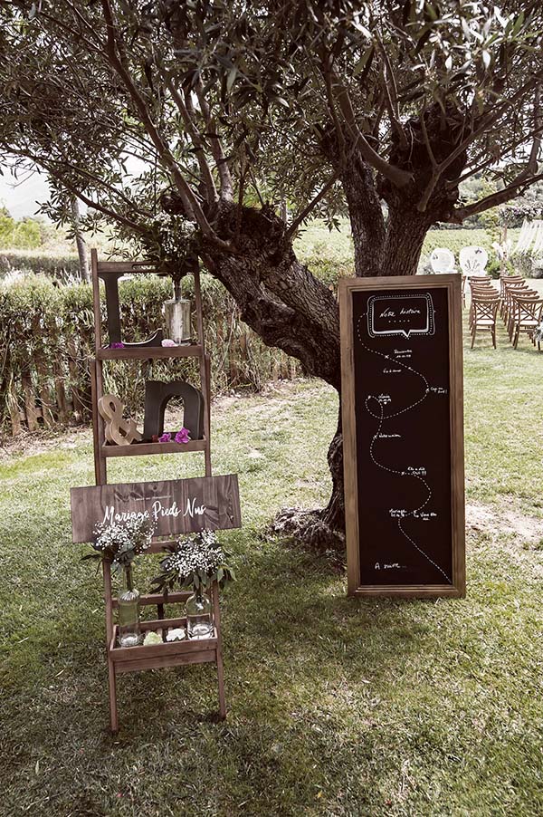 Décoration de mariage champêtre avec échelle en bois, fleurs et plan de table noir sous un olivier.