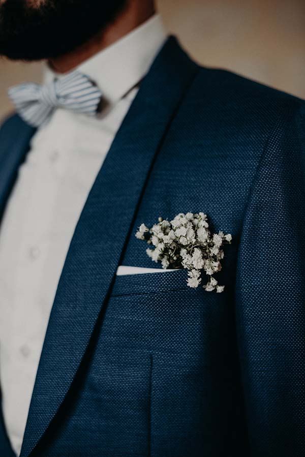 Gros plan d'un marié en costume bleu marine avec un nœud papillon rayé et une pochette fleurie blanche.