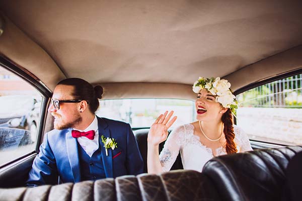 Jeunes mariés souriants à l'arrière d'une voiture ancienne, elle en robe blanche et couronne de fleurs, lui en costume bleu.