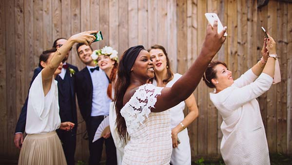 Des invités souriants en tenue de cérémonie prennent des selfies devant un mur en bois dans une ambiance festive.