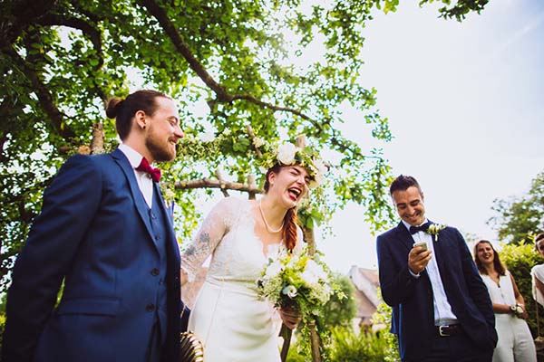 Jeunes mariés riant sous des arbres, elle en robe blanche et couronne de fleurs, lui en costume bleu marine.