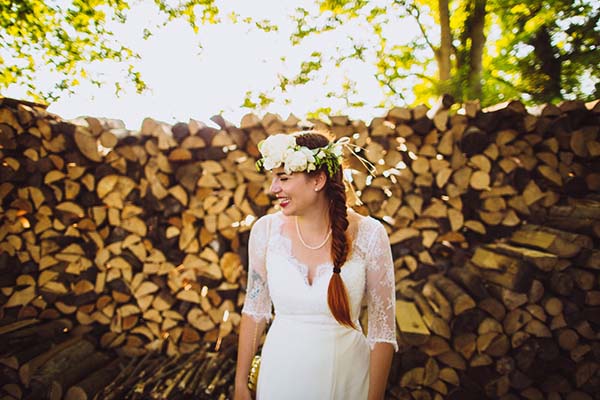 Mariée souriante en robe blanche et couronne de fleurs devant un mur de bois, ambiance bucolique et lumineuse.