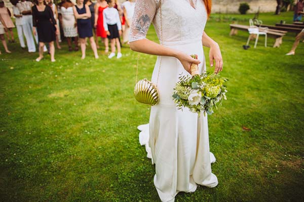 Mariée en robe blanche tenant un bouquet champêtre, avec un petit sac doré, lors d'un mariage bucolique en extérieur.
