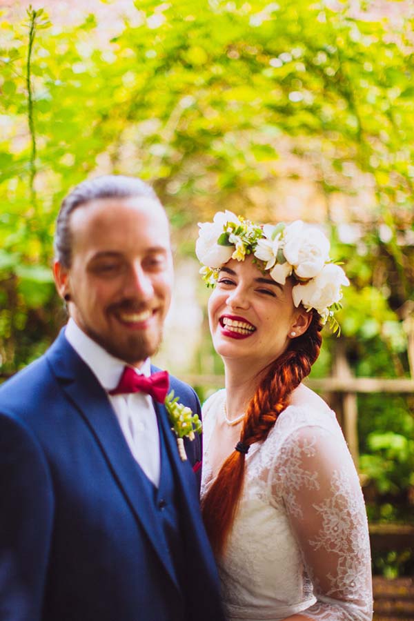 Jeunes mariés souriants en tenue festive, la mariée portant une couronne de fleurs blanches dans un cadre verdoyant.