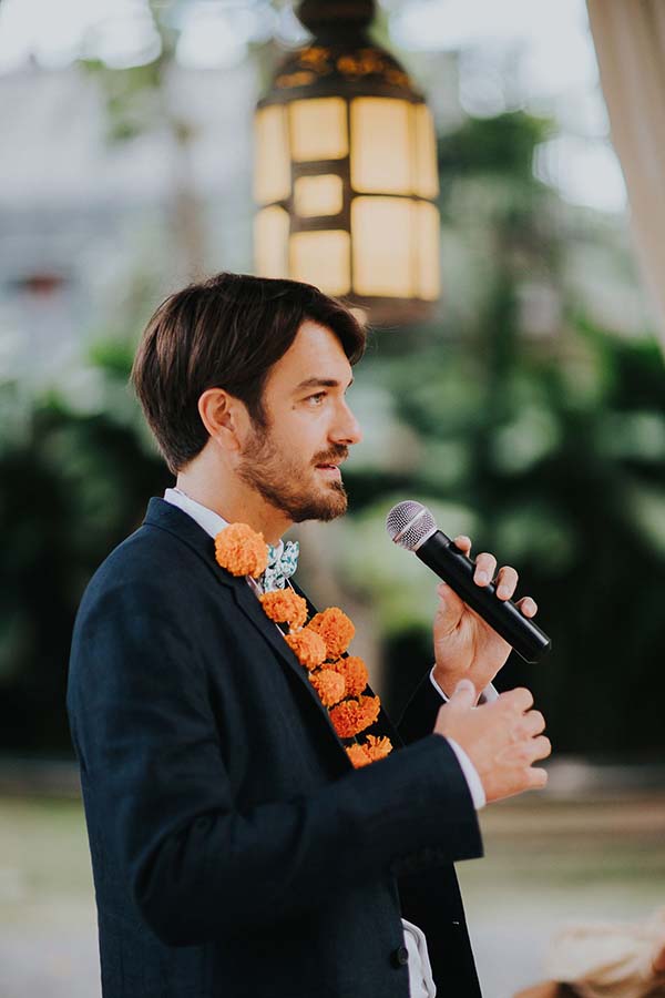 Homme en costume bleu avec un collier de fleurs orange parlant au micro lors d'une cérémonie en extérieur.