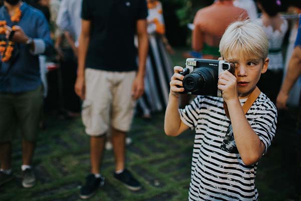 Un jeune garçon blond en t-shirt rayé prend une photo avec un appareil argentique lors d'une fête en extérieur.