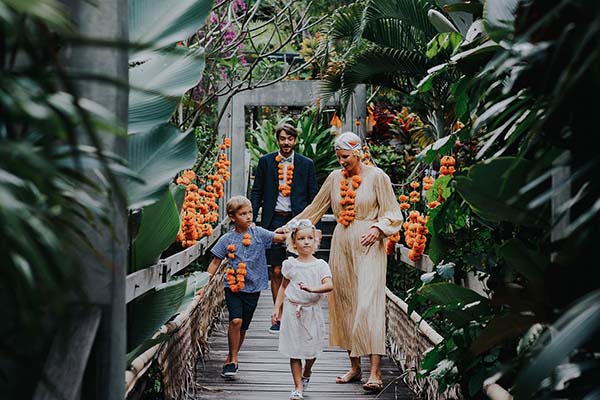 Une famille en tenue élégante et colliers de fleurs marchant sur un pont en bois dans un jardin tropical à Bali.
