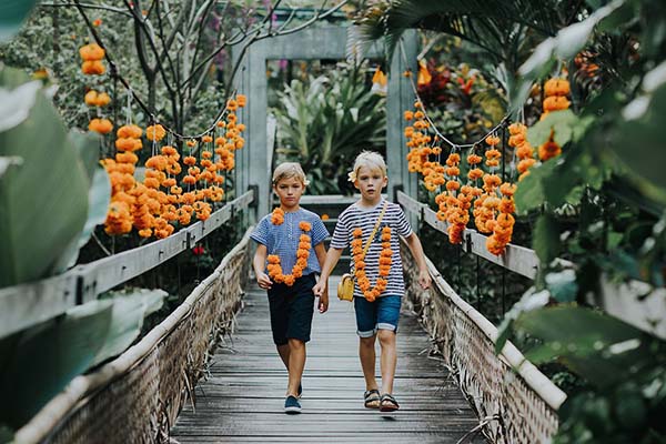 Deux jeunes garçons en tenue estivale marchent sur un pont en bois décoré de fleurs orange dans un jardin tropical.