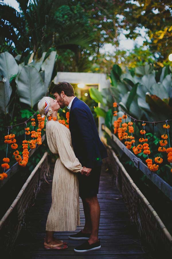 Couple s'embrassant sur un pont en bois décoré de fleurs orange dans un jardin tropical luxuriant à Bali.