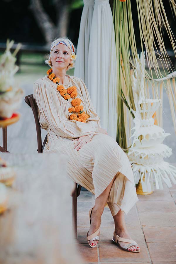 Femme en robe plissée beige et collier de fleurs orange, assise lors d'une cérémonie à Bali.