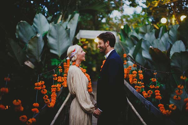 Couple souriant se tenant la main dans un jardin tropical balinais décoré de guirlandes de fleurs orange.