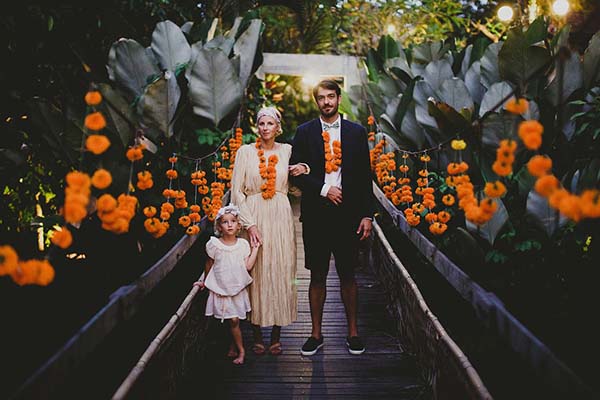 Couple et enfant en tenue blanche et noire posant sur un pont décoré de fleurs orange dans un jardin tropical à Bali.