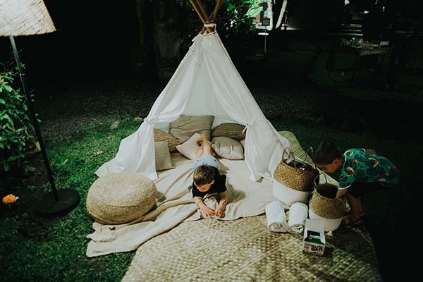 Espace détente pour enfants avec tipi blanc et coussins beiges sur une pelouse nocturne à Bali.