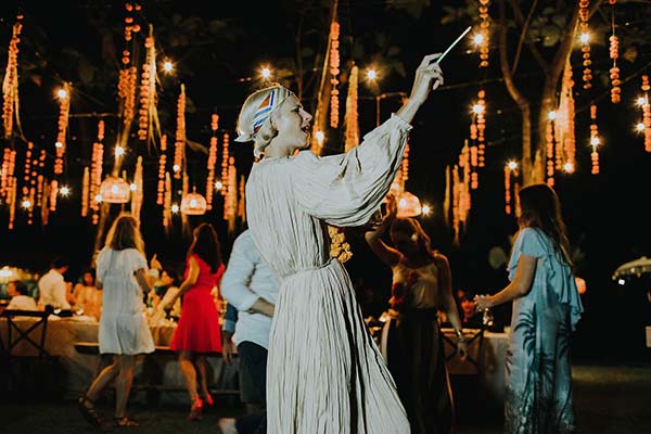 Une femme en robe blanche et coiffe originale dirige un orchestre lors d'une soirée festive et illuminée à Bali.