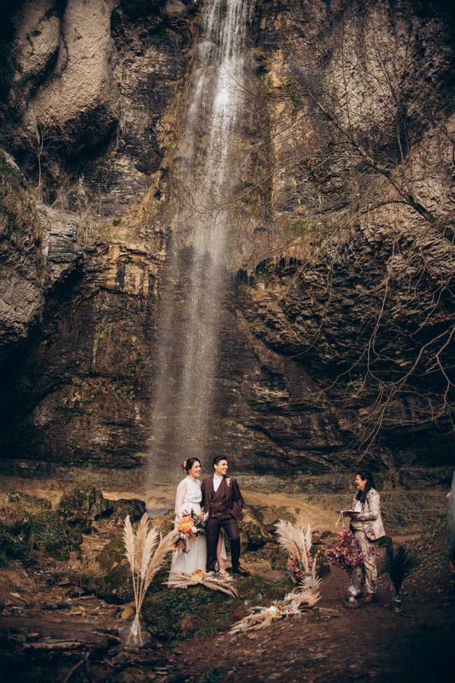 Couple de mariés et officiante lors d'un elopement romantique au pied d'une haute cascade et de falaises rocheuses.
