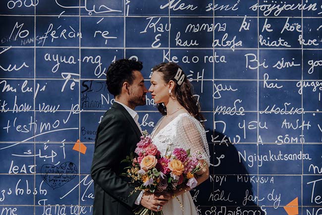 Un couple romantique debout devant un mur bleu orné de phrases amoureuses, célébrant leur amour avec une bouquet coloré.