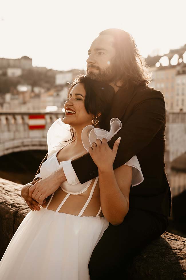 Un couple heureux partage un moment romantique sur un pont urbain ensoleillé, vêtus de vêtements de mariage éclatants.