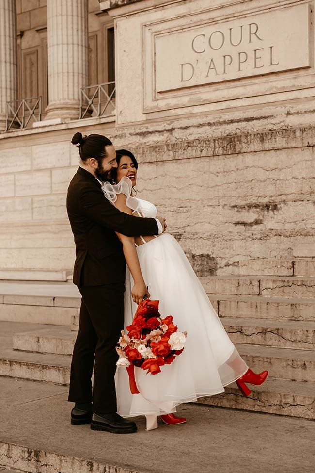 Un couple heureux dans une robe blanche et un costume sombre, tenant un bouquet de fleurs rouges, dans un décor urbain raffiné.
