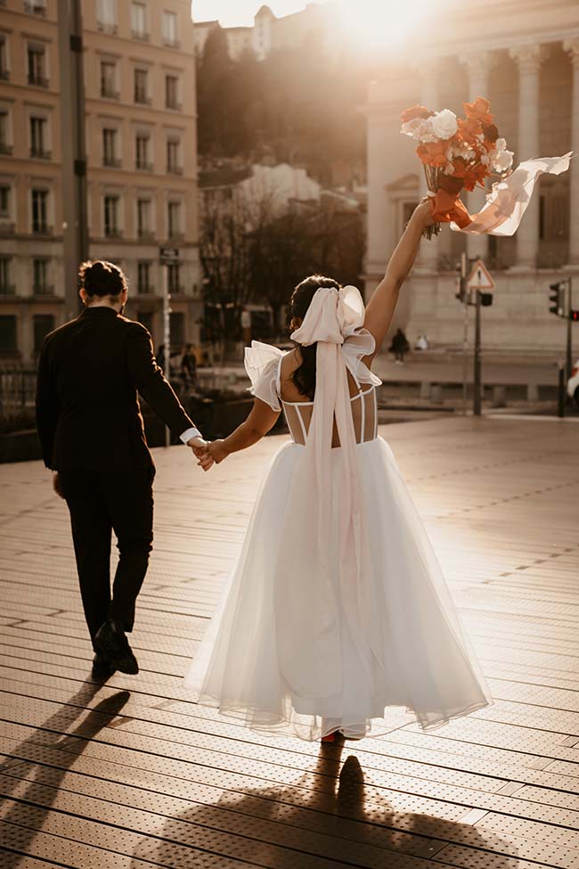 Une mariée en blanc et son mari dans un costume noir dans une scène urbaine ensoleillée, avec des tons chauds et romantiques.