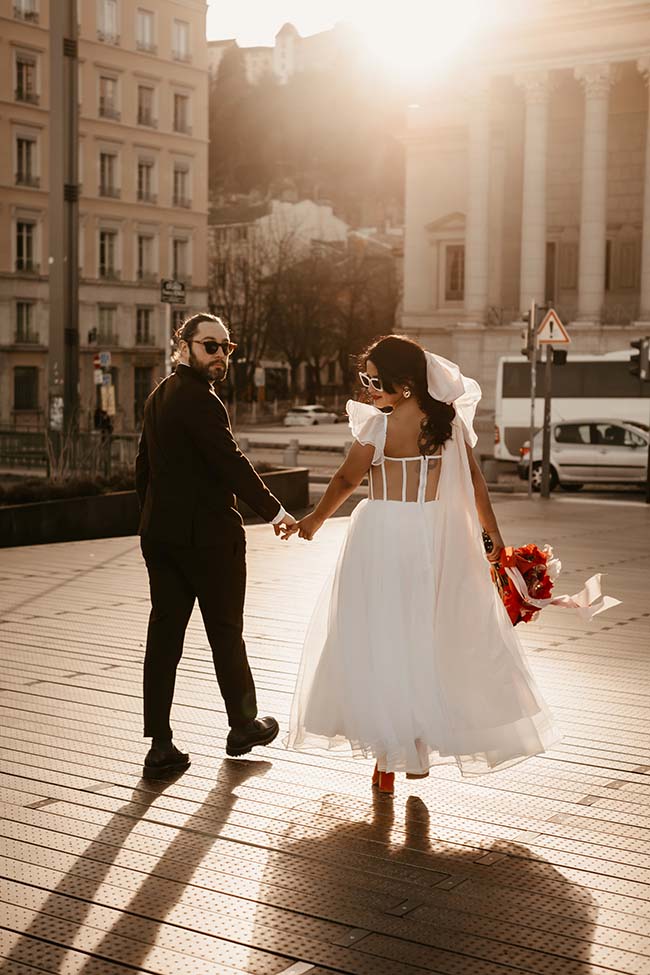Une mariée en robe blanche et un mari en costume sombre se promènent main dans la main sur un pont urbain, éclairés par