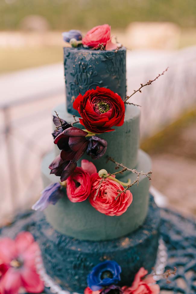Un gâteau de mariage moderne avec une touche de fantaisie, orné de fleurs rouges et noires luxueuses sur un fond vert profond.