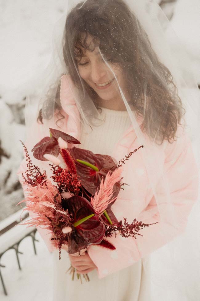 Une mariée sous la neige, habillée d'un doux manteau rose, déguste l'émotion en tenant un bouquet de fleurs vives.