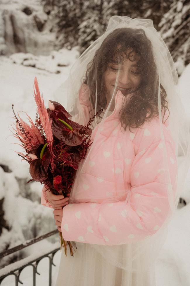 Une mariée charmante dans un manteau rose avec coeur, tenant un bouquet de fleurs sombres, debout dans un décor neigeux.